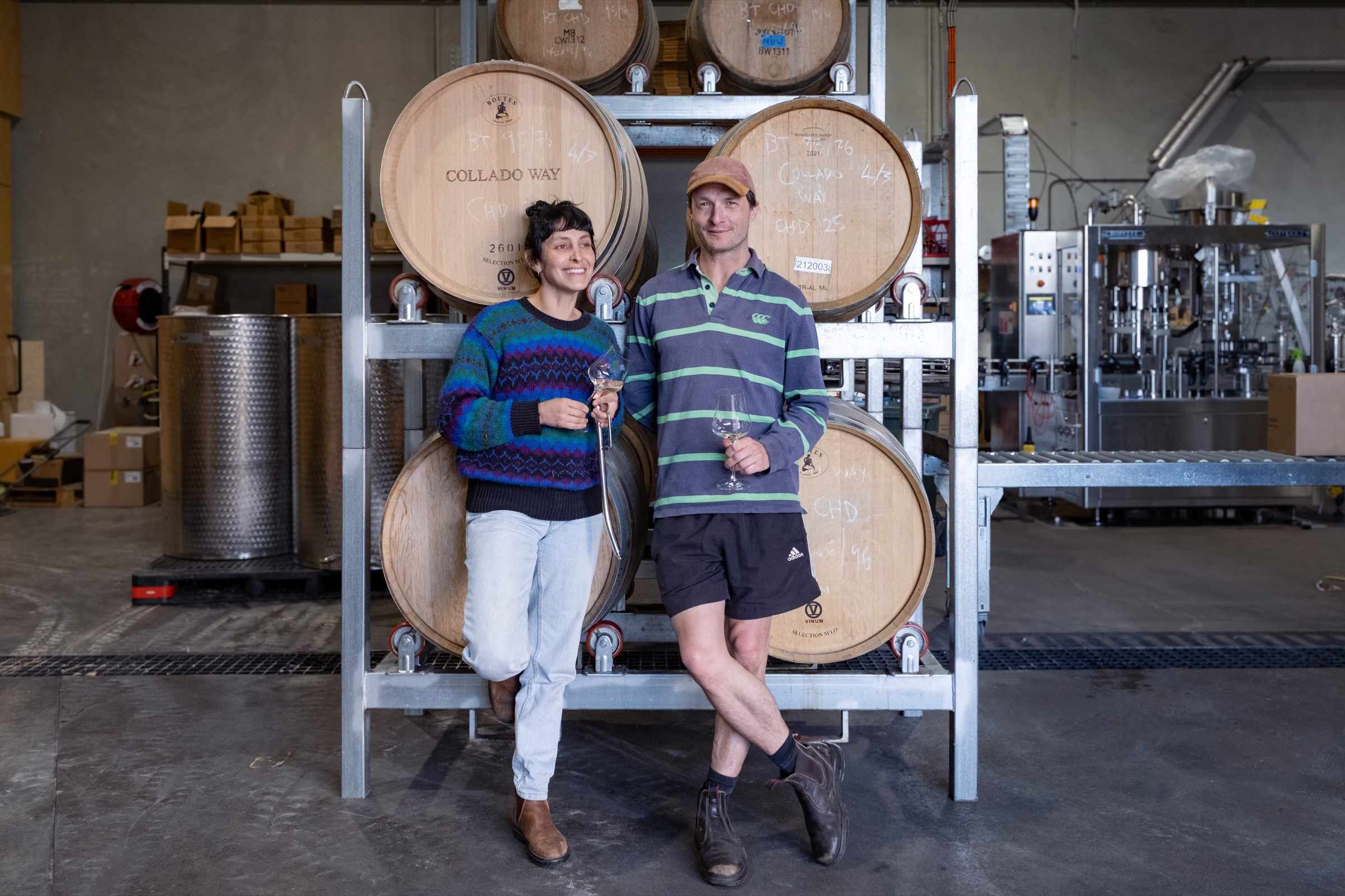 Two people holding glasses of wine in front of barrels in the Collado Way Wines winery